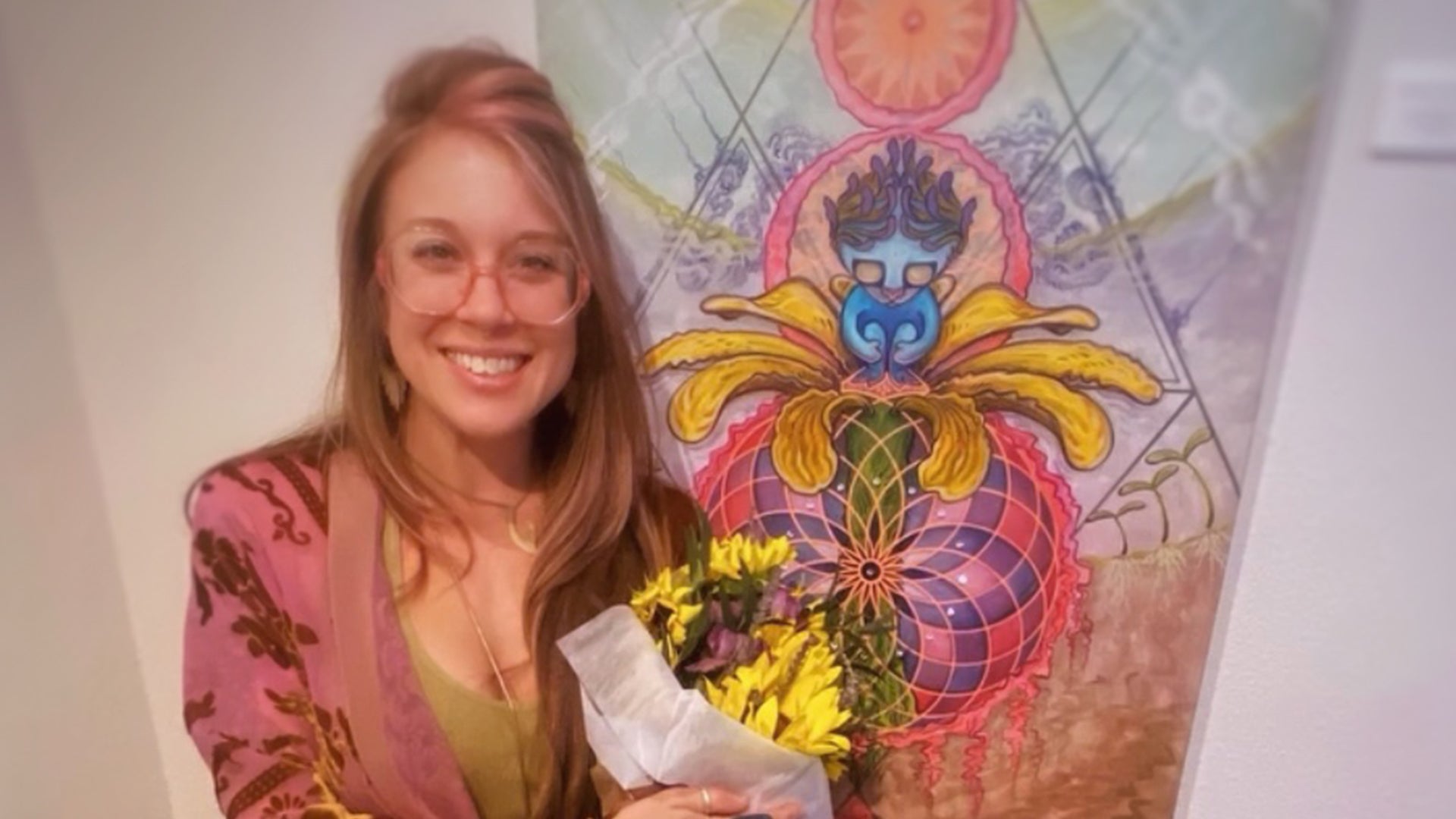Woman with long hair and glasses holds yellow flowers before a vibrant, abstract painting featuring geometric patterns and mystical, colorful imagery.